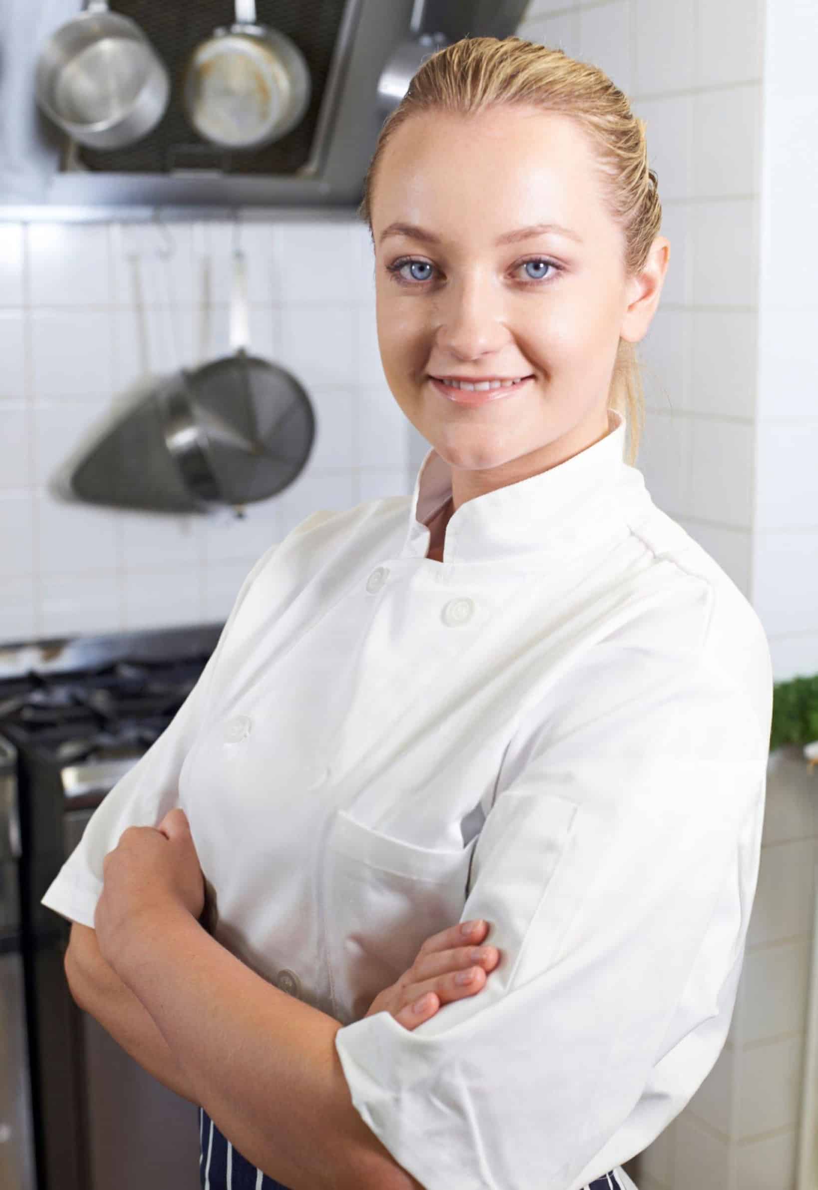 Maya Bennett, Forkful Daily’s Head Chef, smiling confidently in a white kitchen setting, wearing her chef coat with arms folded.