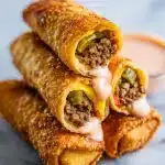 Three golden crispy Big Mac egg rolls with melted cheese, sesame seeds, and a bowl of pink dipping sauce on a wooden board.