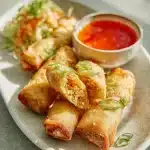 Golden crispy chicken egg rolls arranged on a white plate with a small bowl of vibrant red dipping sauce, garnished with green onions.