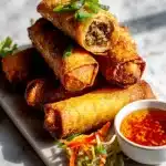 Golden crispy ground beef egg rolls on a marble surface with glossy dipping sauce, fresh scallions, and colorful slaw.