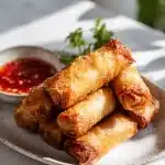 Golden crispy pork egg rolls with savory filling, on a plate garnished with green onions, beside small bowl of sweet chili dipping sauce.