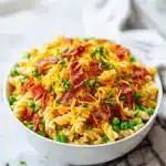 A vibrant, creamy Bacon Ranch Pasta Salad loaded with golden crispy bacon, shredded cheddar, bright green peas, and fresh herbs, glistening under natural light.