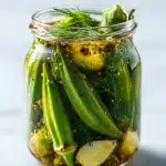 A glass mason jar filled with vibrant bright green pickled okra pods, whole garlic cloves, and fresh dill sprigs, submerged in a clear, spice-flecked brine on a rustic wooden surface.