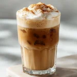 A tall glass of iced banana cloud latte with layers of golden espresso and creamy banana cold foam, topped with a sprinkle of cinnamon, on a white marble countertop.