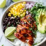 A vibrant BBQ chicken rice bowl with smoky glazed chicken strips, black beans, corn, avocado, fresh cilantro, red onion, and lime wedge.