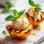 Two grilled peach halves caramelized to golden, topped with melting vanilla ice cream, fresh mint, and honey drizzle, a bbq peaches recipe.
