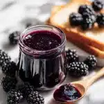 A jar of glossy deep purple homemade blackberry jam with fresh blackberries, a wooden spoon, and toast on a rustic table in natural light.