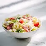 A colorful bow tie pasta salad with salami, mozzarella, cherry tomatoes, bell peppers, and basil in a white bowl on a rustic wooden table, bright natural light.