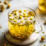 Mug of golden chamomile sleep tea with floating chamomile blossoms, honey dipper, lemon slice, and scattered dried lavender buds on a dark wood table.