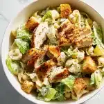 A vibrant bowl of Chicken Caesar Pasta Salad with juicy chicken, romaine, croutons, creamy dressing, and shaved Parmesan.