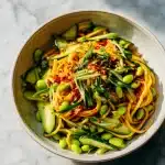 A bowl of Cold Sesame Noodles with Cucumber & Edamame topped with scallions and chili oil on a bright marble surface.