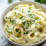 Creamy cottage cheese alfredo sauce coating fettuccine, topped with fresh parsley and grated Parmesan, on white marble.