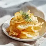 Crispy golden-brown cottage cheese chips with black pepper and fresh dill garnish, arranged on a white plate over linen. Airy and light texture visible.