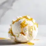 A round scoop of white cottage cheese ice cream with glossy golden honey drizzled on top, sitting on a bright white marble surface.