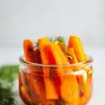 Vibrant orange fermented carrot sticks with fresh dill and garlic in clear brine in a glass jar, on a bright clean background.