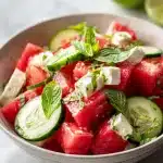 Vibrant bowl of feta cucumber watermelon salad with watermelon cubes, fresh mint and basil, lime dressing on light marble.
