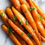 A skillet filled with glossy, caramelized garlic brown sugar carrots, garnished with bright green fresh parsley. The carrots are tender and vibrant, with a rich, sticky glaze coating each piece.