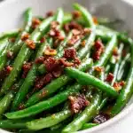 A close-up of sautéed garlic brown sugar green beans in a glossy butter sauce, topped with crispy bacon crumbles, served in a cast-iron skillet on a rustic wooden board with steam rising.