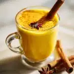 A steaming mug of golden milk bedtime drink with a cinnamon stick and star anise, on a marble counter in soft natural light.