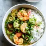 A steaming white bowl of gumbo recipe with okra and shrimp, topped with fresh scallions and paprika, with fluffy rice beside it.