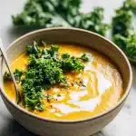 Bowl of gut friendly lentil stew with fresh kale, a swirl of coconut milk, and red lentils visible on a light marble surface.
