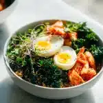 A beautifully plated gut friendly quinoa bowl with vibrant kimchi, sautéed kale, a halved soft-boiled egg, and fresh microgreens on a light background.