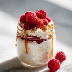 Glass jar filled with layers of high fiber overnight oats including creamy yogurt-soaked rolled oats, plump red raspberries, speckled chia seeds, and a glossy maple syrup drizzle, placed on a marble surface with a spoon beside it.
