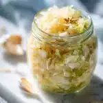 A glass jar filled with glistening, pale shredded fermented cabbage, speckled with visible caraway seeds and whole garlic cloves, sitting on a rustic wooden surface with a soft, natural light highlighting the crunchy texture of the homemade sauerkraut.