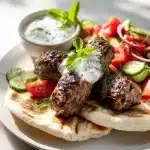 Close-up of juicy kefta kebabs from a kefta kebab recipe with warm pita, tomato-cucumber salad, and yogurt sauce, natural light.