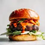 A towering kimchi burger on a brioche bun, with red gochujang mayo, charred beef patty, crisp kimchi, and lettuce on a bright clean surface.