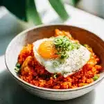 Vibrant red kimchi fried rice in a steaming bowl, topped with a glossy fried egg, fresh green onions, and sesame seeds, on a rustic table.