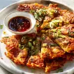 Close-up of crispy golden kimchi pancake with scallions, visible tangy kimchi pieces, and a bowl of savory dipping sauce.
