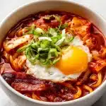 A steaming bowl of kimchi udon with glossy red sauce, crispy bacon, fresh green onions, and a vibrant egg yolk.