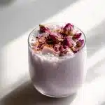 A glass mug of creamy purple lavender moon milk with dried rose petals and a cinnamon stick on a wooden table.