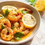 A cast iron skillet with lemon garlic butter shrimp, garnished with fresh parsley and lemon wedges on a wooden board.
