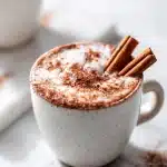 A steaming mug of magnesium hot chocolate for sleep topped with frothy milk, cocoa powder dust, and a cinnamon stick on a light background.