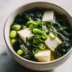 A steaming bowl of miso soup for gut health with silken tofu, wakame, edamame, and bok choy, garnished with green onions and ginger.