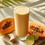 A creamy papaya digestive smoothie in a tall glass, with a lime garnish, fresh papaya and ginger on a marble surface.