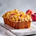 A slice of moist peach bread with a golden cinnamon crumb topping, resting on a white plate with fresh peach slices and a dusting of powdered sugar, on a wooden table.