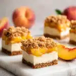 Square dessert bars with visible layers of buttery graham cracker crust, thick creamy cheesecake, and a golden brown cobbler crumble topping studded with fresh peach slices, arranged on a bright white marble surface.