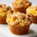 Golden-brown peach cobbler muffins with cracked streusel and juicy peach chunks on a white marble surface under soft light.