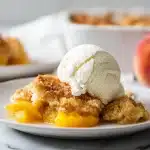 Peach cobbler with cake mix in a white dish with golden crust and vanilla ice cream