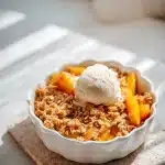Rustic peach crisp with golden oat crumble topping and melting vanilla ice cream on a bright stone surface.