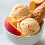 Two scoops of creamy homemade peach ice cream in a white ceramic bowl with fresh peach wedges and a waffle cone piece on a light marble surface.