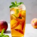 Refreshing homemade peach iced tea in a tall glass, garnished with fresh peach slices and mint, glowing in natural light.