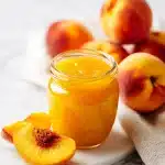 Golden peach jam in a mason jar, next to a cut fresh peach and a slice of toasted bread smeared with jam, on a rustic wooden surface.