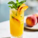 A tall crystal-clear glass filled with vibrant orange peach lemonade, garnished with fresh peach slices and a sprig of mint on a light background.