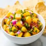 A white bowl filled with fresh peach salsa, featuring diced peaches, red onion, cilantro, and jalapeño, with tortilla chips around.