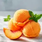 Creamy scoops of vibrant orange peach sorbet garnished with fresh peach wedges and mint leaves on a bright marble surface, a perfect summer dessert.