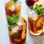 A tall glass of iced peach tea with fresh peach slices, mint leaves, and ice cubes, on a wooden table with a rustic backdrop.